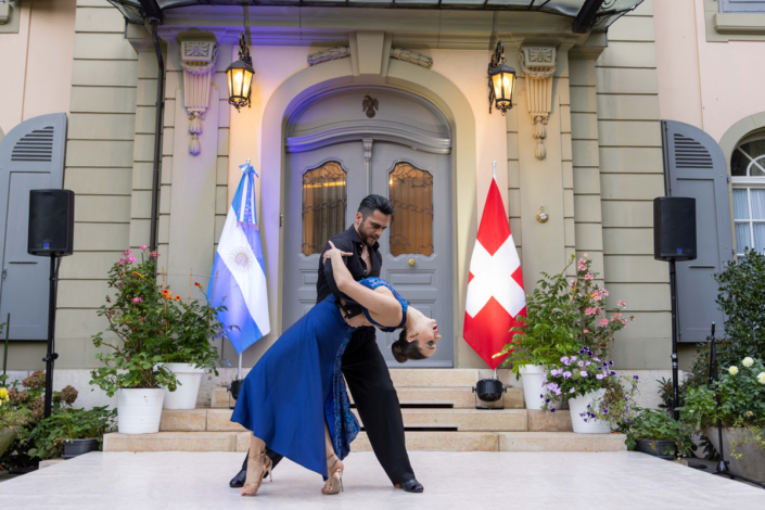 A couple dances in front of a house with Swiss flags.