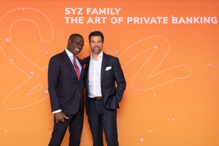 Zwei Männer in Anzügen stehen neben einem orangefarbenen Hintergrund mit der Aufschrift „Sy2 Family – Die Kunst des Private Banking“.