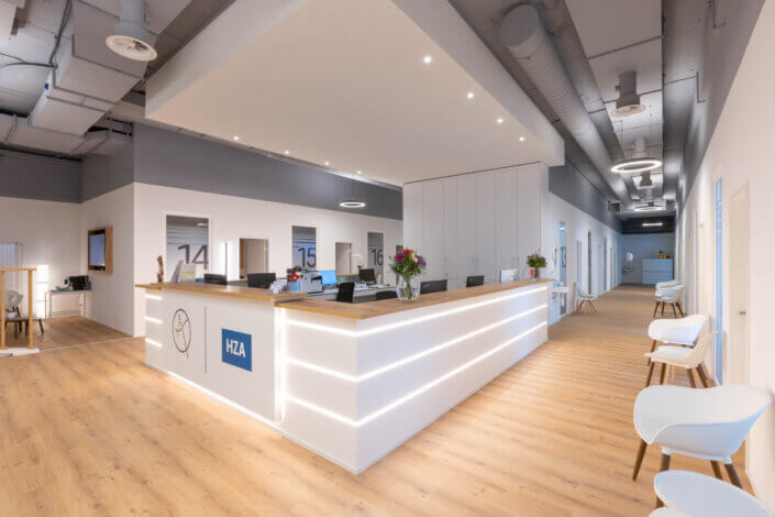 Reception area in a modern design with a long white desk, wooden floor, chairs and ceiling lighting. The walls have numbered rooms and there are exposed cables on the ceiling.