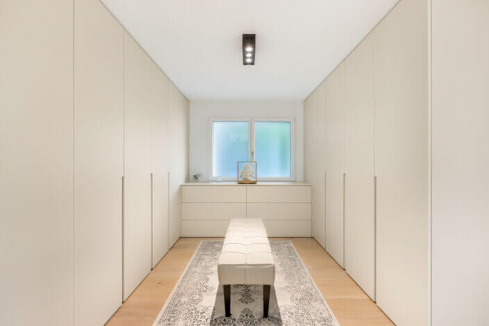 Minimalist walk-in closet with white cupboards, a white upholstered bench in the middle, a rug and a window with frosted glass for natural light.