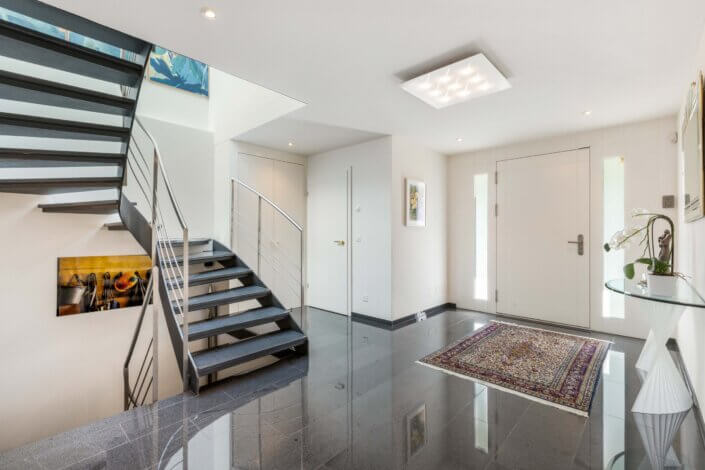 Modern entrance area with shiny black tiled floor, a patterned carpet and a metal staircase. Works of art hang on the white walls. There is a door to the outside on the right-hand side.