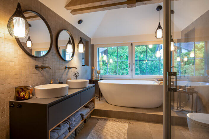 Modern bathroom with two round mirrors above a double washbasin, a free-standing bathtub next to large windows and pendant lights. Neutral tones and wooden accents enhance the room.