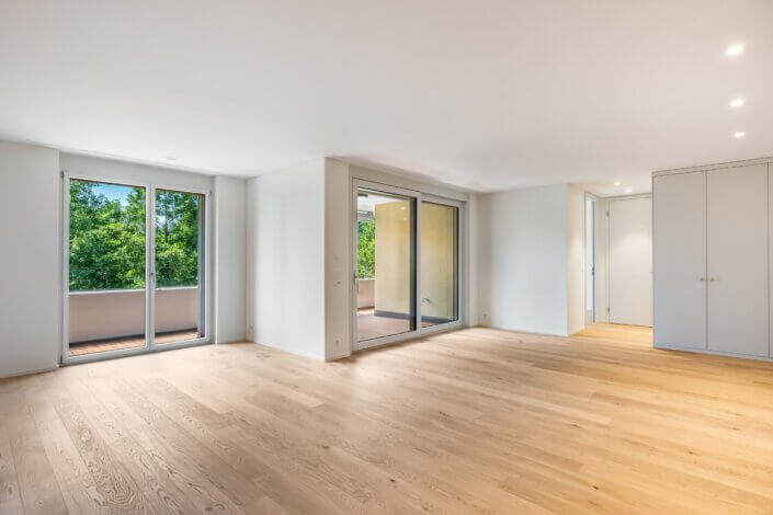 Empty room with a light wooden floor, large windows and sliding glass doors leading onto a balcony. The walls are white and there are closed cupboard doors on the right-hand side.