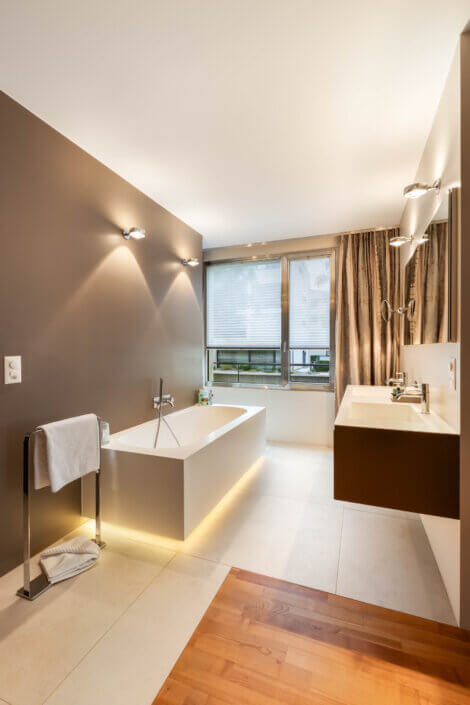 Modern bathroom with brown accent wall, bathtub, double washbasin and large window with blinds. The floor is a mixture of wood and tiles. Wall lights illuminate the room.