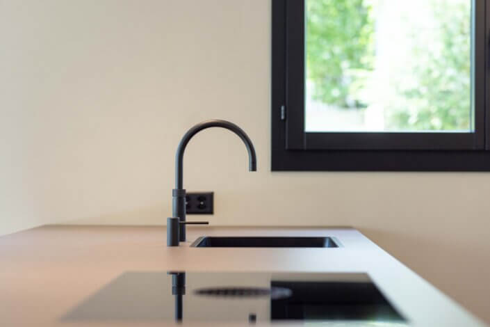 Minimalist kitchen with black tap, sink and induction hob on a light-colored worktop. There is a window in the background that lets in natural light.