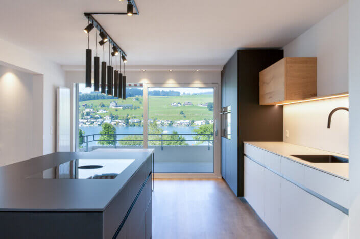 Modern kitchen with elegant black and white cabinets, cooking island and hanging lamps. Large windows offer a view of a lake and green hills in the background.