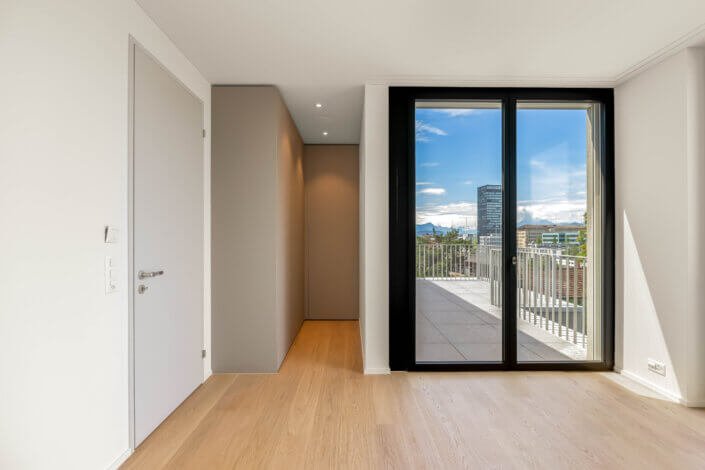 Minimalist room with wooden floor, white walls and a glass door leading onto a balcony with a view of the city. On the left is a closed white door.