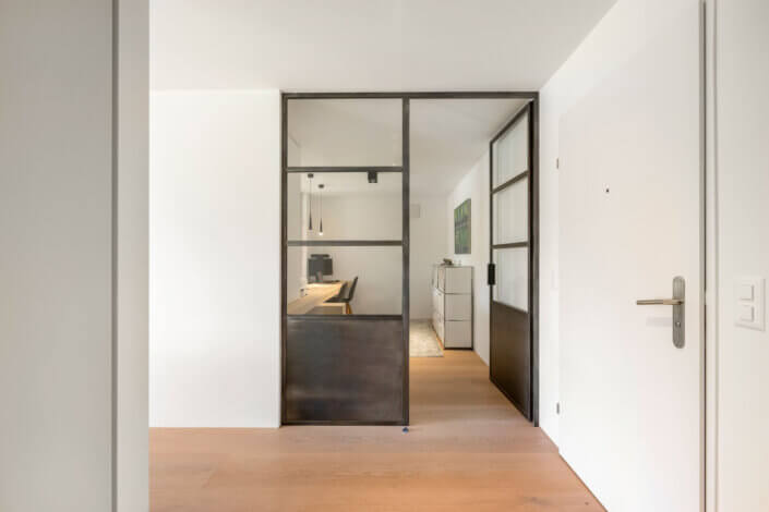 A glass door with a black frame opens into a modern office space with a wooden floor, a large desk and two chairs. White walls and a white door can be seen in the foreground.