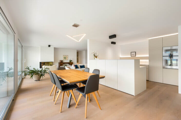 Modern open-plan interior with a wooden dining table and black chairs, a minimalist kitchen island, plants and large windows.