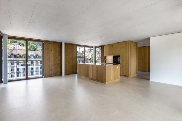 Spacious, modern kitchen and living area with large windows, light wooden cupboards and views of the buildings and the green surroundings outside.