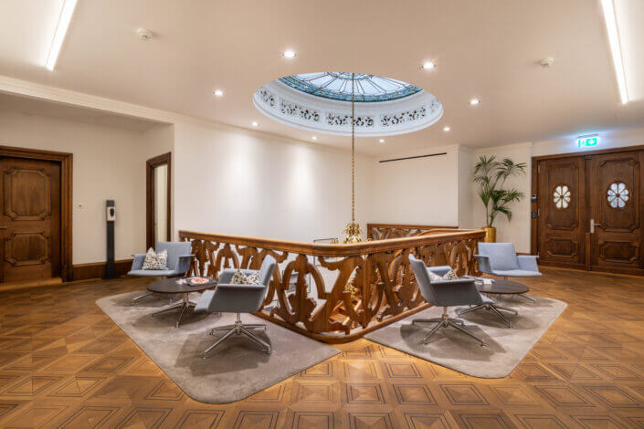 A modern lounge area with four swivel chairs around a wooden railing under a decorative skylight. Wooden doors and a potted plant can be seen in the background.