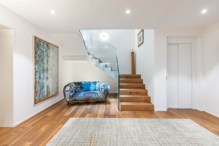 Spacious room with wooden staircase, glass railing and modern sofa. Large abstract painting on the wall and textured carpet on the wooden floor.