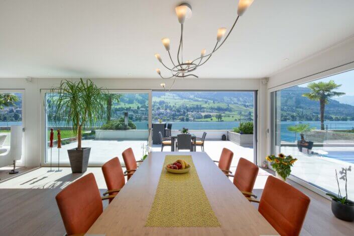 Modern dining room with wooden table, orange chairs and yellow runner. Large windows offer lake and mountain views. Elegant chandelier above, with plants and views of the outdoor terrace.