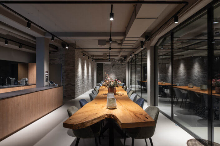 Modern conference room with a long wooden table surrounded by chairs. The room has exposed brick walls, ceiling lights and glass partitions that create an industrial and elegant atmosphere.