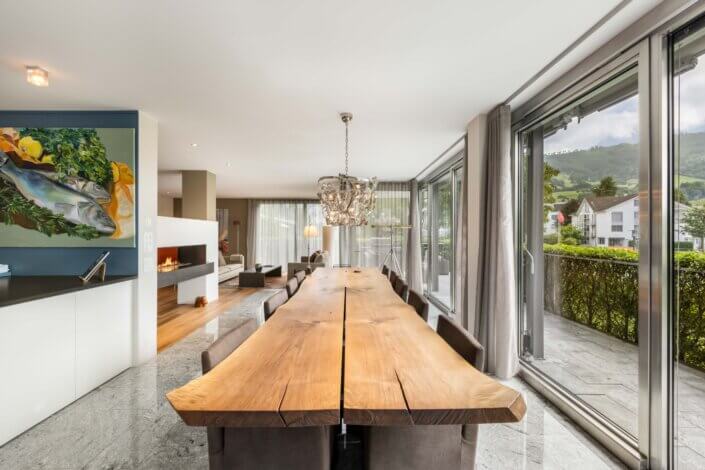 Modern dining room with a long wooden table, black chairs, large windows and a view of the garden and mountains. A contemporary work of art hangs on the wall and a chandelier sits enthroned above the table.