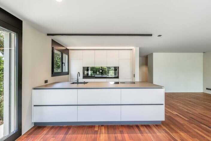 Modern, minimalist kitchen with elegant white cabinets, built-in appliances, large kitchen island and wooden floor. There is a large window on the left-hand side that provides natural light.