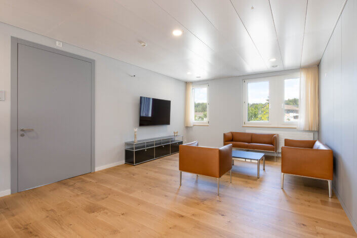 Minimalist living room with two brown leather armchairs, a matching sofa, a wall-mounted TV and a modern glass coffee table. Light wooden floor and large windows with beige curtains.