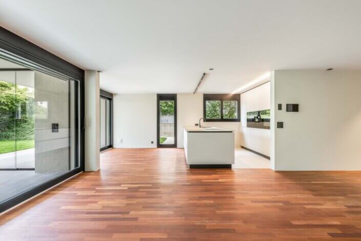 Modern, open interior with wooden floor, large windows and a central kitchen island. The room is bright and offers views of a garden through glass doors.