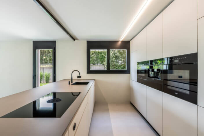 Modern kitchen with white cabinets, black worktops, built-in appliances and a large window. The room has a minimalist design with clean lines and natural light.