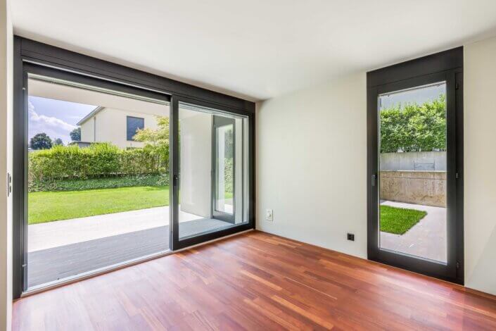 Empty room with large windows and wooden floor, leading onto a grassy backyard with a concrete patio. The walls are white and the room is bright thanks to the natural light.