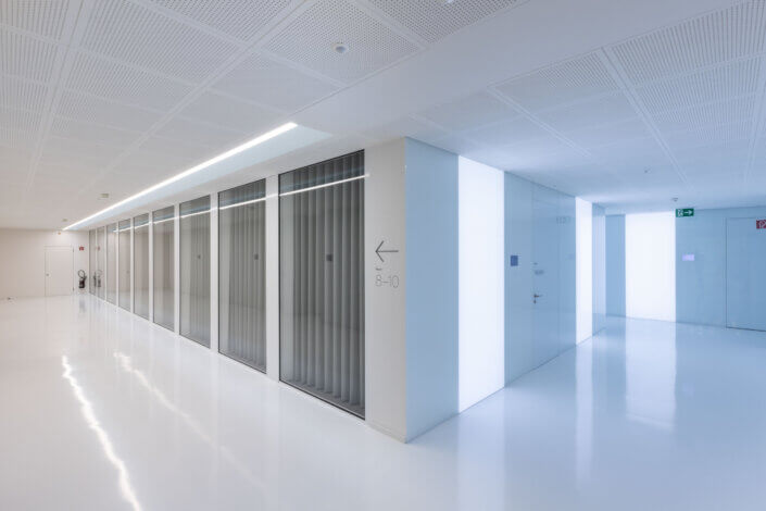 A modern, bright and minimalist hallway with white walls and a reflective floor. One side is lined with glass partitions and a small sign points the way.