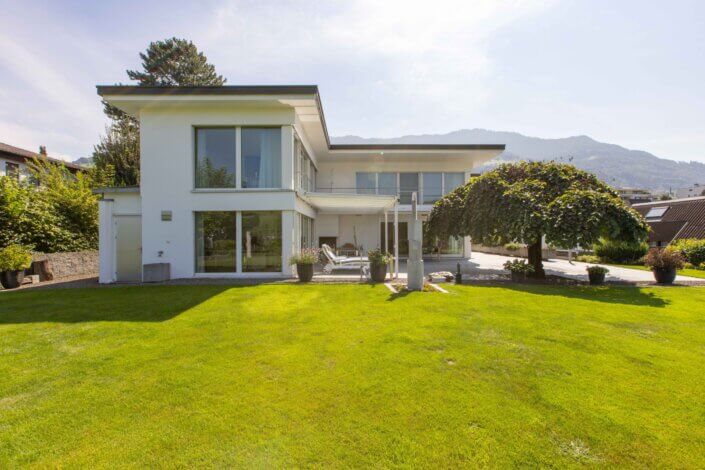 Modern two-story white house with large windows and manicured lawn against a backdrop of hills and trees.