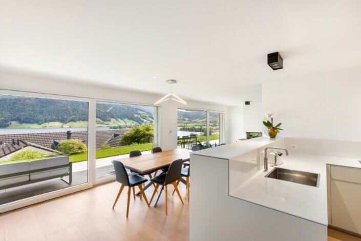 Modern open-plan kitchen and dining area with views of the mountains and lake through large glass doors leading onto a terrace. White worktops and a wooden table with chairs can be seen.