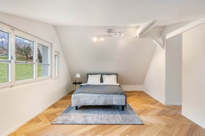 Minimalist bedroom with a double bed on a gray carpet, flanked by a small table and lamp. Wooden floor in herringbone pattern, white walls and three windows on the left with a view of the greenery.