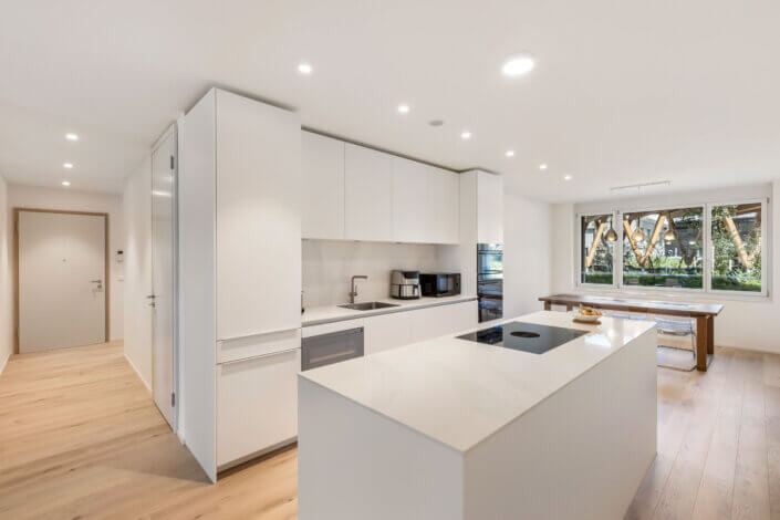 Modern kitchen with white cupboards, a cooking island and a dining area with wooden table. Large window offers a view of the greenery. Light wooden floors and recessed lighting are available.