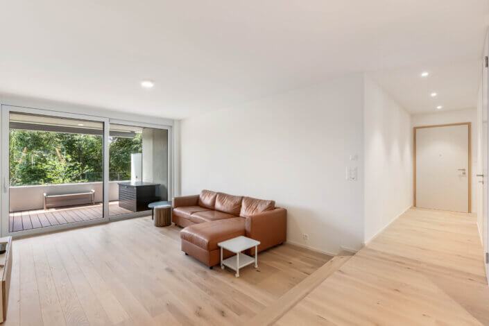 Spacious living room with a brown leather sofa, a small white table and sliding glass doors leading onto a balcony with lots of greenery. Light wooden floor and white walls everywhere.