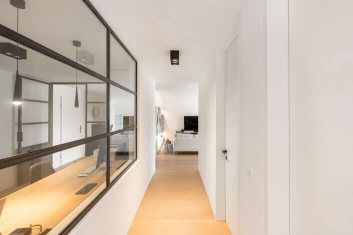 Minimalist hallway with wooden floor and white walls. On the left is a glass partition wall that separates a work area with a desk. At the end is a living area with sofa and TV.