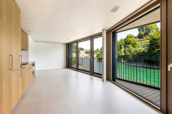 Modern, unfurnished room with large windows overlooking a river and trees, with light-colored wooden cabinets and concrete floor.