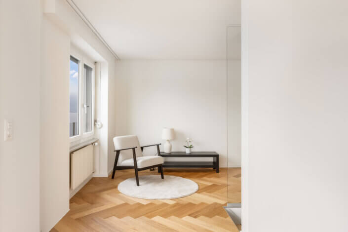 Minimalist room with wooden chair, white seat upholstery, round carpet and small table with lamp and plant. Wooden floor with chevron pattern and a window with a view outside.