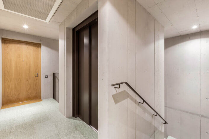 Hallway with elevator doors, a wooden door on the left, concrete walls and a staircase with metal railing. Bright lighting from ceiling lights.