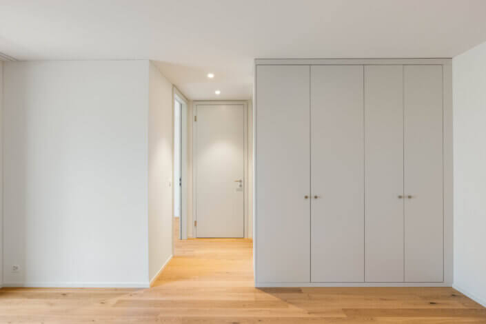 Minimalist interior with a light wooden floor, white walls, a closed white closet, recessed lighting and a door leading into another room.