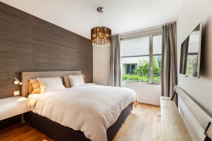 A modern bedroom with a large bed, white bed linen, bedside lamps, a wall-mounted TV and a window with curtains. There is a decorative ceiling light and wooden floor.