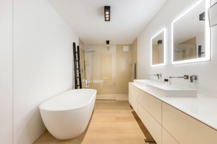 Modern bathroom with free-standing bathtub, double washbasin with illuminated mirrors, glazed shower and light wooden floor.