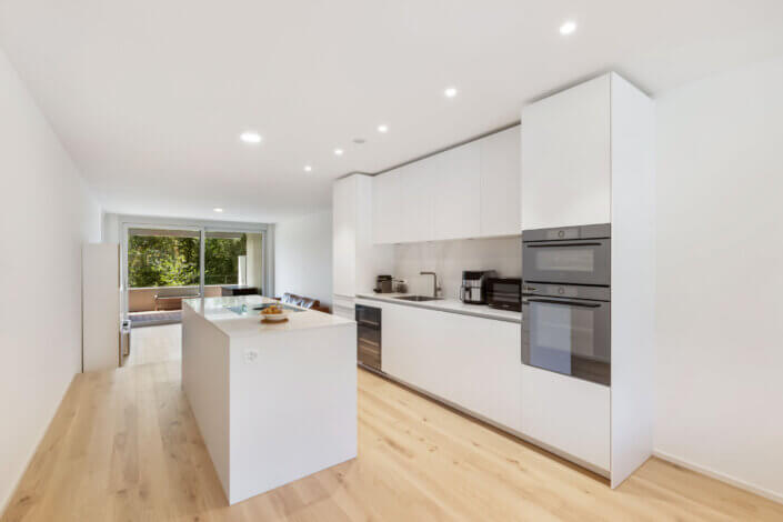 Modern kitchen with white cabinets, built-in double ovens and a cooking island. The wooden floor extends into a living area with a glass door leading to an outdoor area with lots of greenery.